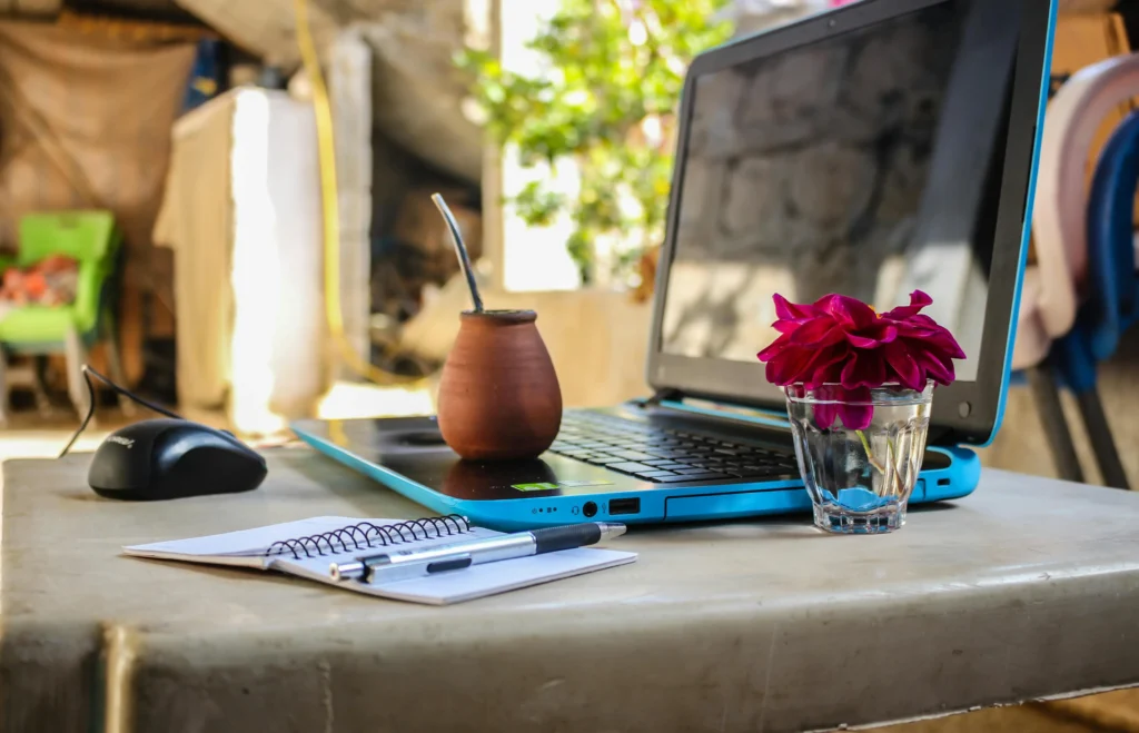 A laptop and a notebook on the table with a flower and drink.