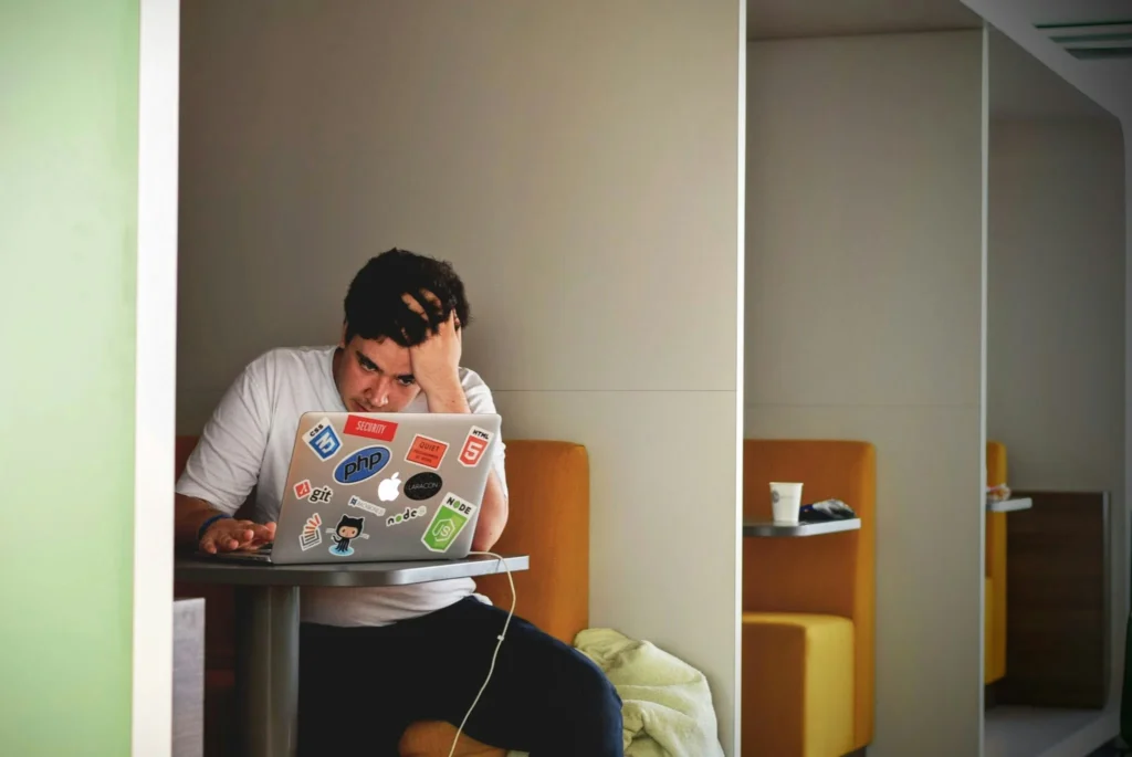 A person rubbing their head while working on a laptop in a booth as sign of entrepreneurial isolation