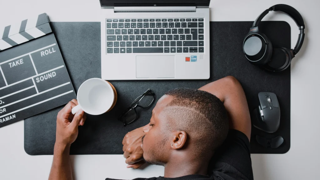 A man sleeping at their desk while holding a cup, surrounded by a laptop and tech items.